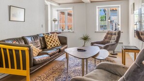 Una sala moderna con un sofá de cuero, un sillón a juego, una pequeña mesa de centro y un banco de madera. Hay dos ventanas que proporcionan luz natural y vistas al exterior.
