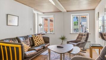 Una sala moderna con un sofá de cuero, un sillón a juego, una pequeña mesa de centro y un banco de madera. Hay dos ventanas que proporcionan luz natural y vistas al exterior.