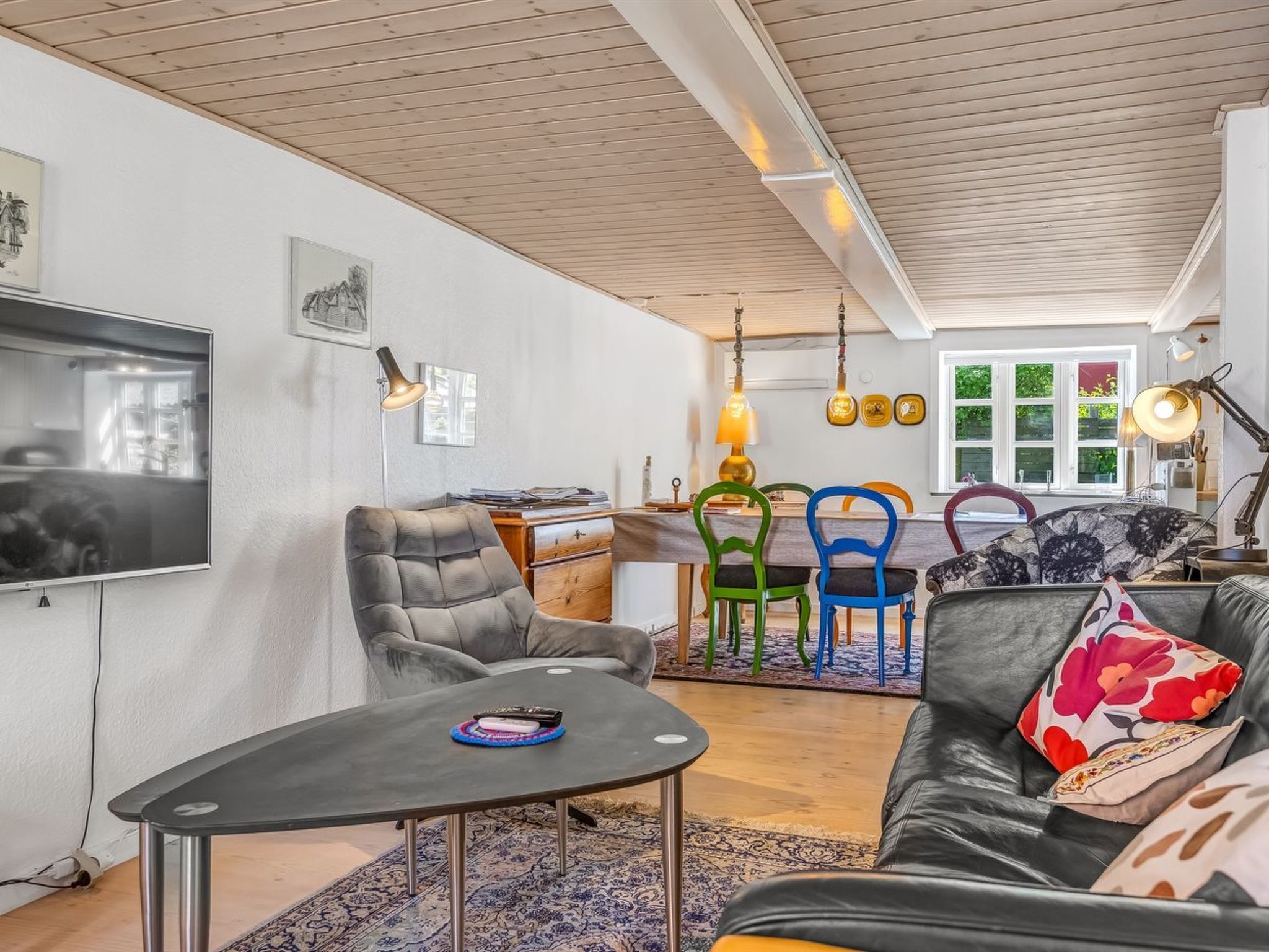 Una sala de estar moderna con un televisor, un área de comedor con sillas coloridas y un sofá con cojines estampados.