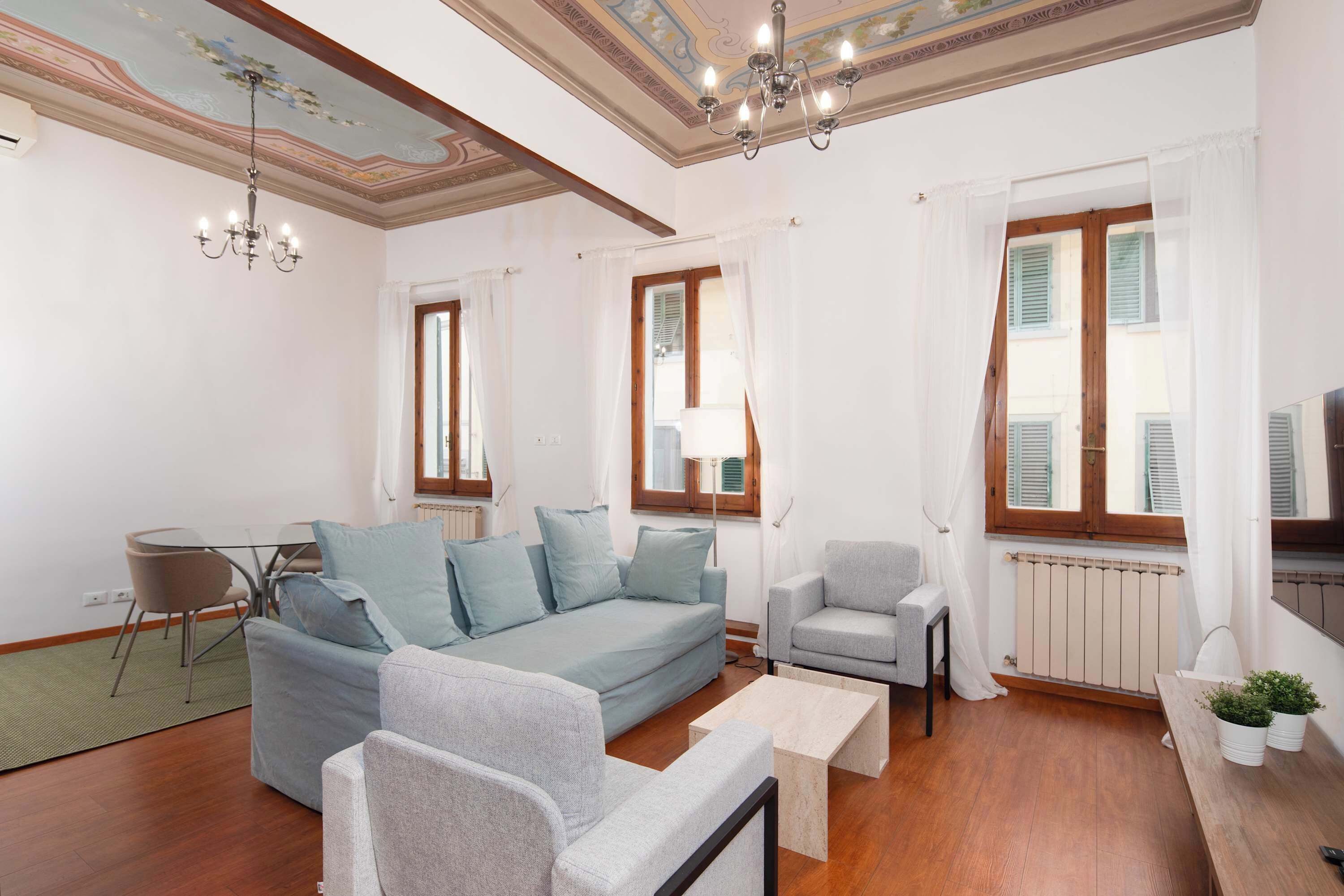 Una sala de estar con sofá, sillones, una mesa de centro y una mesa de comedor. Hay ventanas con contraventanas, un candelabro y un radiador.
