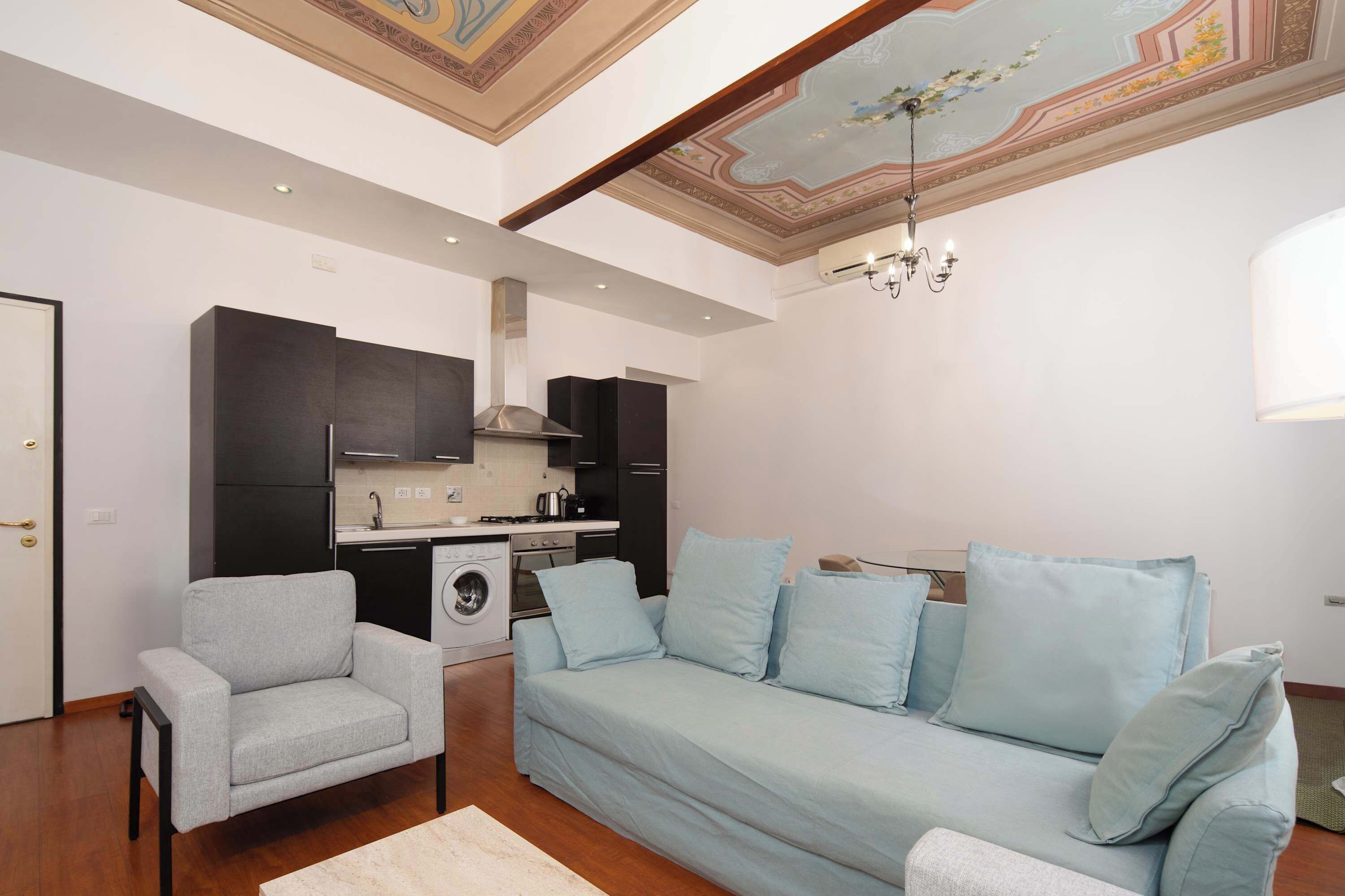 A compact living space with a kitchenette, a sofa, a chair, and a chandelier.