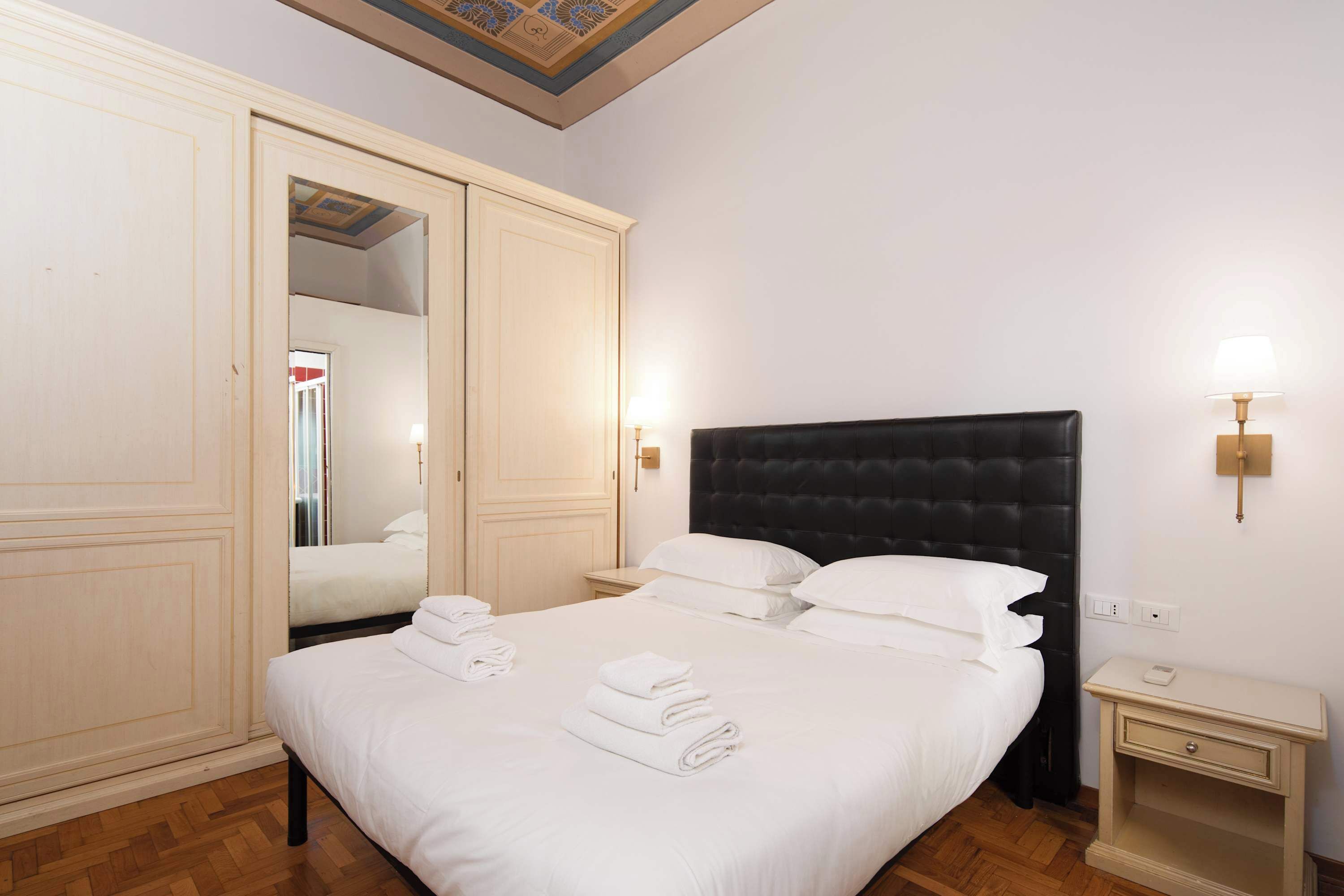 A hotel room with a large bed, a wardrobe, a nightstand, and a wall-mounted lamp.