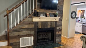 A living room with a fireplace, a staircase, a TV mounted above the fireplace, and a kitchen in the background. - Cozy Pickwick Lake house overlooking Safe Harbor Aqua Yacht Marina Sleeps 10+ (Iuka)