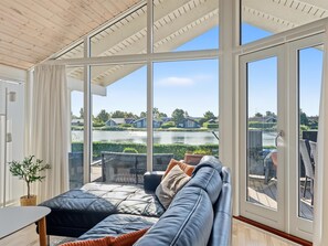 A modern living room with a large glass wall, a sofa, and a view of a lake and houses. - "Østmar" - 1.3km from the sea by Interhome (Væggerløse)