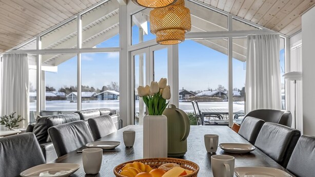 Een eetruimte met een grote tafel, stoelen en uitzicht op een besneeuwd landschap door grote ramen.