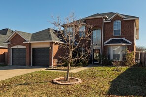 A two-story brick house with a garage, a small tree in the front yard, and a clear sky. - Fire Pit, Kids Toys + Fenced Yard - Family Getaway (Grand Prairie)
