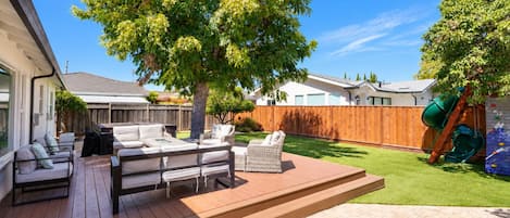 A backyard with a wooden deck, outdoor furniture, a tree, and a play area.