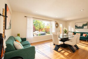 Un salon spacieux avec un canapé vert, une table à manger avec des chaises et une vue sur un jardin à travers de grandes fenêtres.