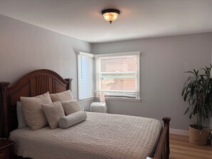 A bedroom with a wooden bed, a nightstand, a bedside table, a potted plant, and a window with blinds. - Belmont Heights 2-bedroom. Quick Walk to 2nd St and Beach (Long Beach)