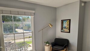 Una habitación con un ventanal grande, una lámpara de pie, una silla y un cuadro enmarcado en la pared.
