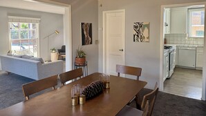 Un comedor con mesa de madera, sillas y ventilador de techo. Hay una puerta que conduce a una cocina y una sala de estar con sofá y lámpara.