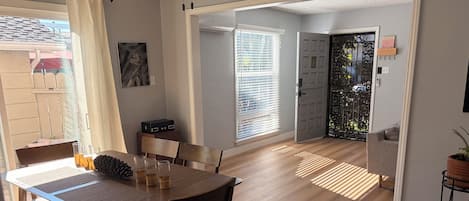 A dining area with a wooden table and chairs, a painting on the wall, and a view of the outside.