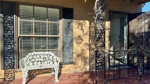 Un patio con un banco de hierro forjado blanco, una reja de metal y un edificio con ventanas y contraventanas grandes.