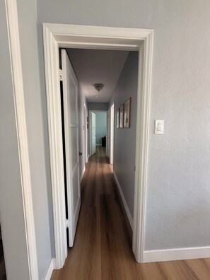 Un pasillo con suelo de madera, paredes blancas y una puerta que da a otra habitación.
