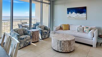 Un vaste espace de vie avec un balcon donnant sur une plage, aménagé avec un canapé blanc, deux fauteuils à motifs et une table basse ronde.