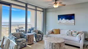 A spacious living area with a balcony overlooking a beach, furnished with a white sofa, two patterned armchairs, and a round coffee table. - Palisades Palms Tw1012 - Sea Escape (Galveston)