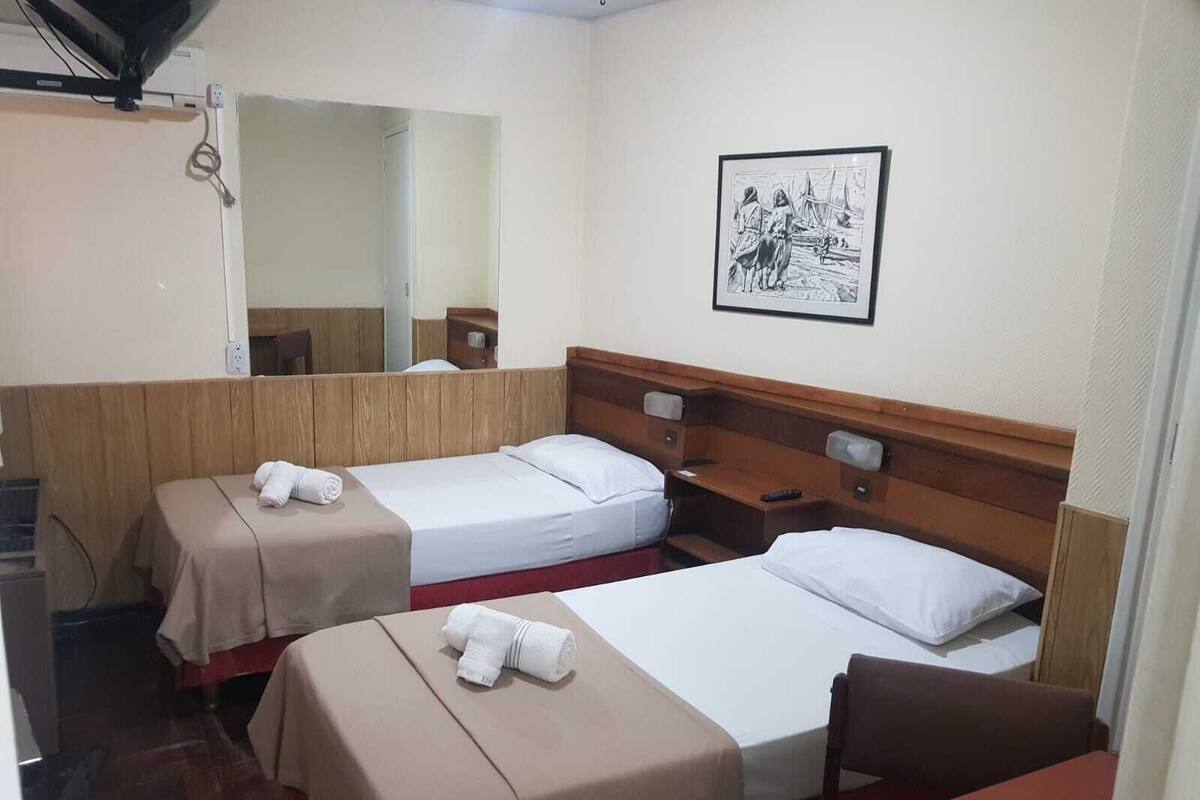 A hotel room with two beds, a wooden headboard, a bedside table, a chair, a desk, a mirror, and a framed picture on the wall.