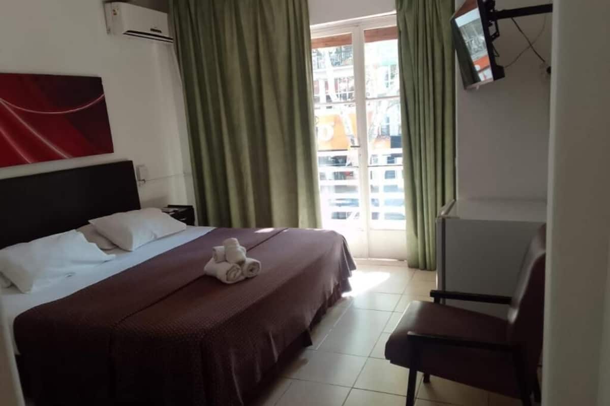 A hotel room with a bed, a chair, a TV, and a window with curtains.