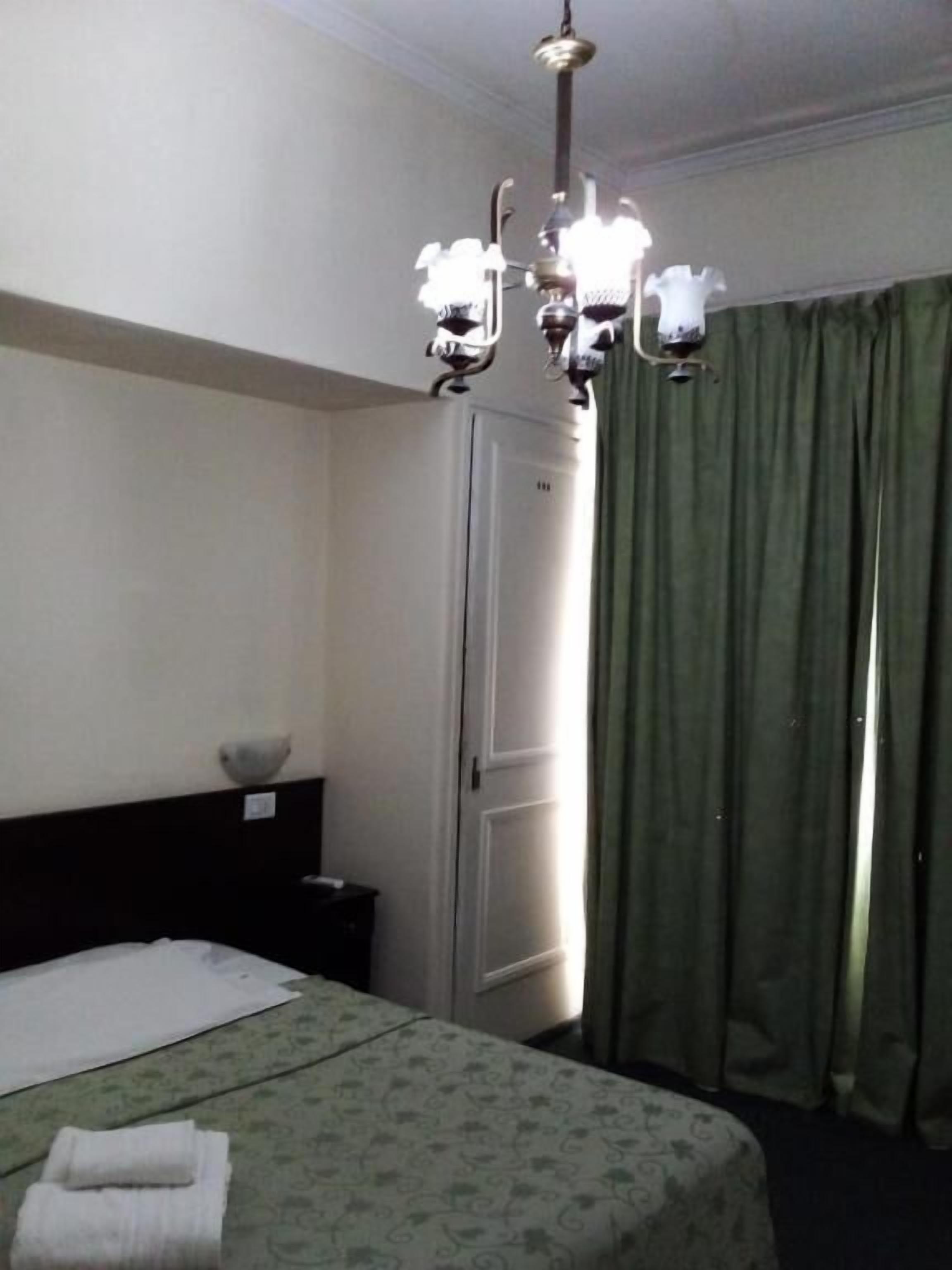 Quarto de hotel com cama, mesa de cabeceira, lustre, porta e cortinas verdes.