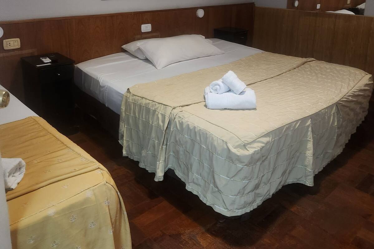 A hotel room with a large bed, a smaller bed, and a desk with a chair. There are two paintings on the wall and a mirror above the bed.
