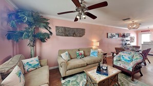 A living room with a beige sofa, a coffee table with a tray, a rattan chair, a ceiling fan, and a large plant. - Sandy Seashell (Port Aransas)