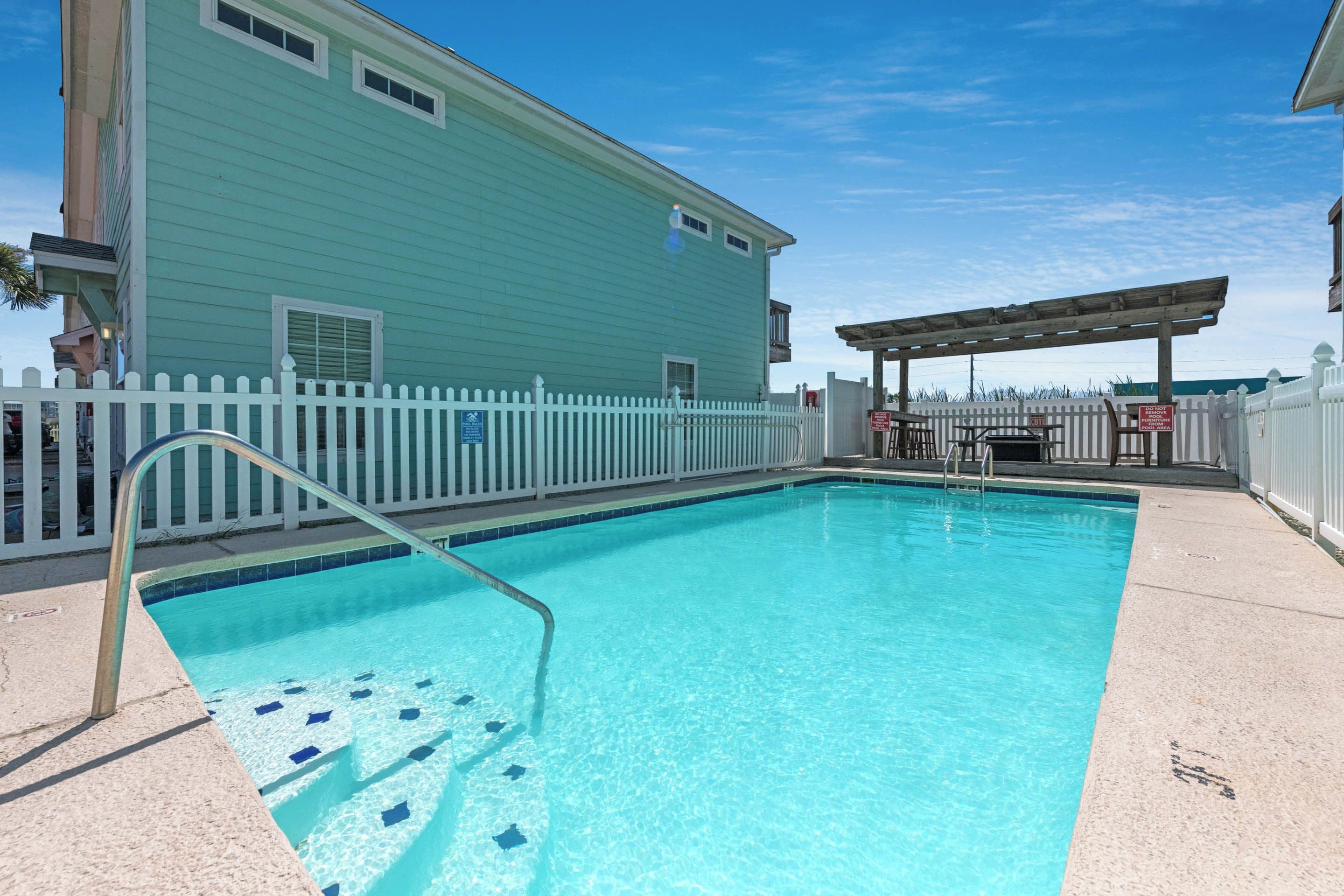 A swimming pool with a white picket fence, a covered patio area, and a light blue building with a balcony.