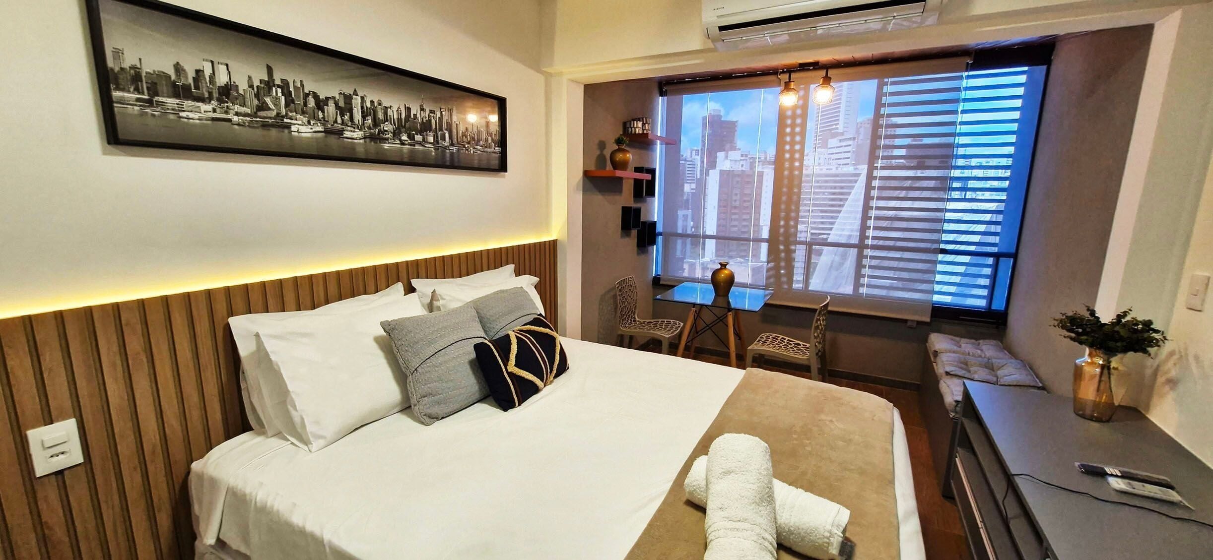 A hotel room with a bed, a desk, a chair, a window with blinds, a cityscape photo on the wall, and a plant on the desk.