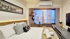 A hotel room with a bed, a TV, a desk with a chair, and a window with blinds. - COBAR0144 - Edifício Único Barra (Salvador)