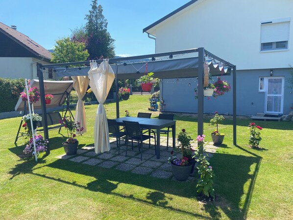 Un patio cubierto con mesa y sillas, rodeado de plantas en macetas y césped.