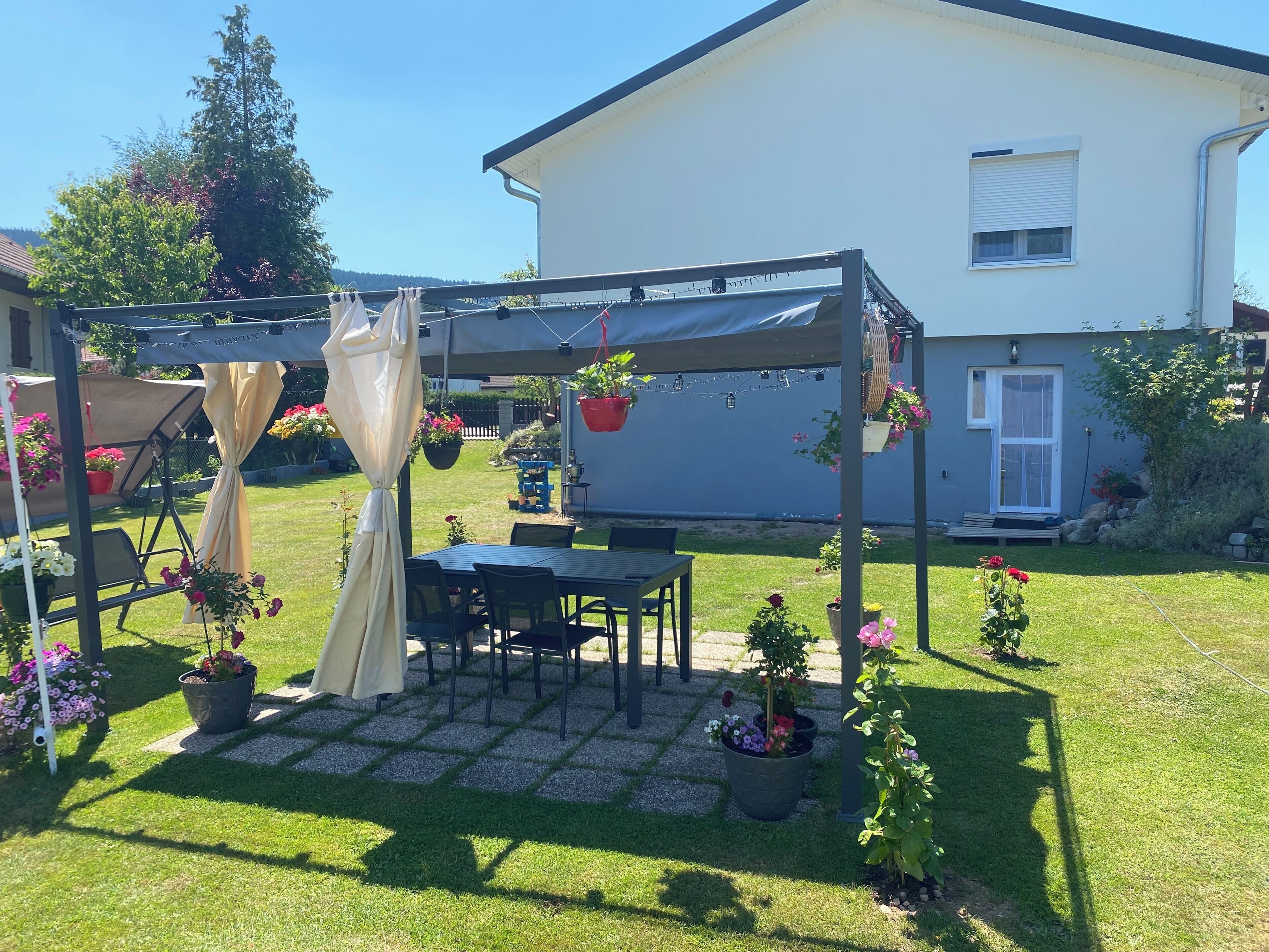 Eine überdachte Terrasse mit Tisch und Stühlen, umgeben von Topfpflanzen und hängenden Blumenkörben.