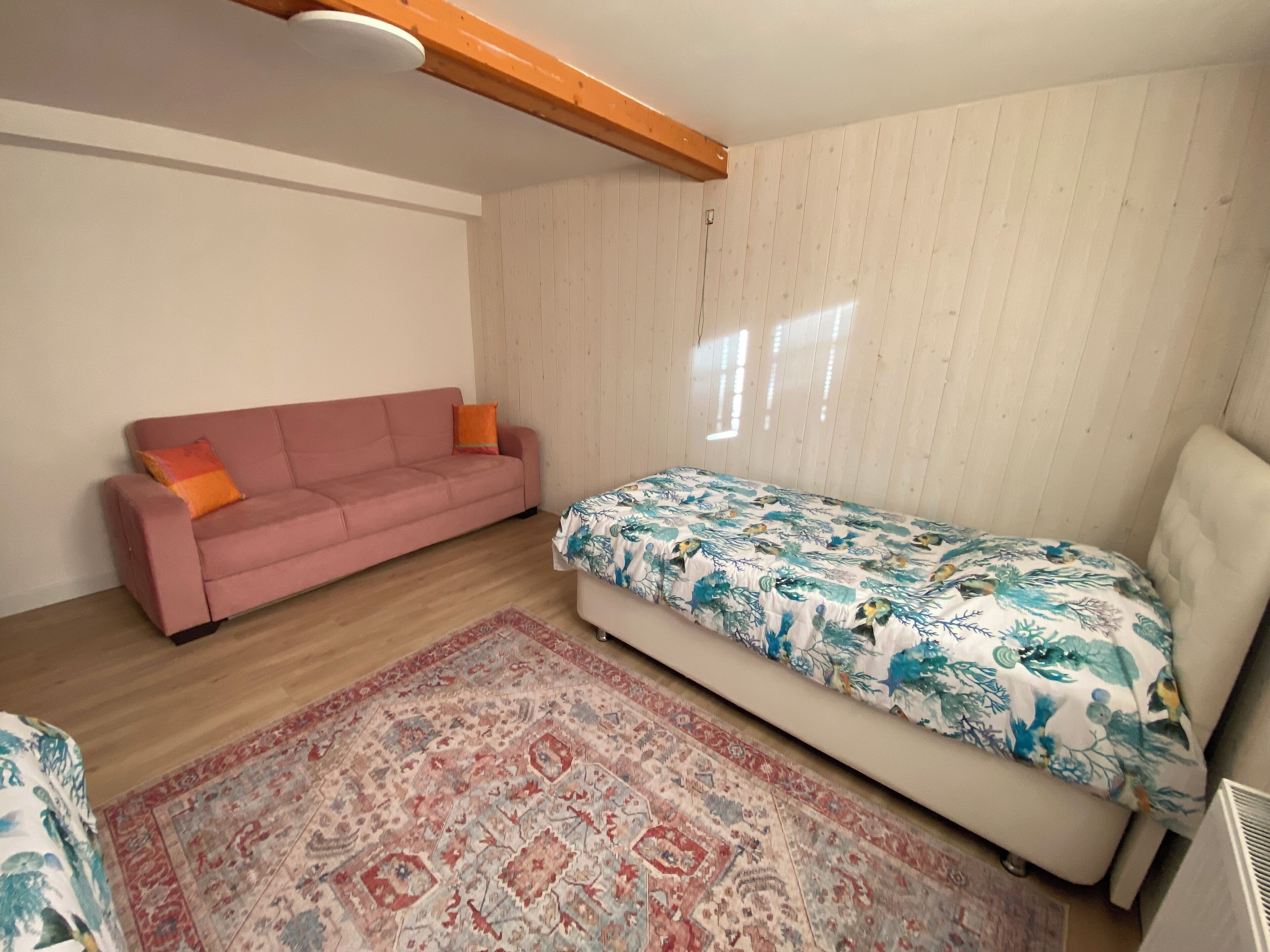 Une petite pièce accueillante, avec un canapé rose, un lit recouvert d’une courtepointe à motifs floraux et un tapis à dessins.