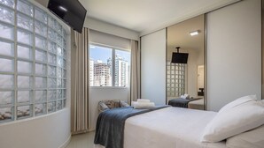 Een moderne hotelkamer met een groot raam, een flatscreen televisie, een bed met witte lakens en een spiegel die de kamer weerspiegelt.