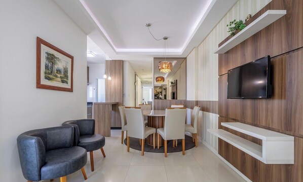 Una sala de estar moderna con sofá, mesa de comedor, sillas, televisor y una cocina al fondo.