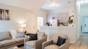 Un salon comprenant un canapé, deux fauteuils, une table basse et un téléviseur. Il y a également un coin cuisine avec des placards et un réfrigérateur.