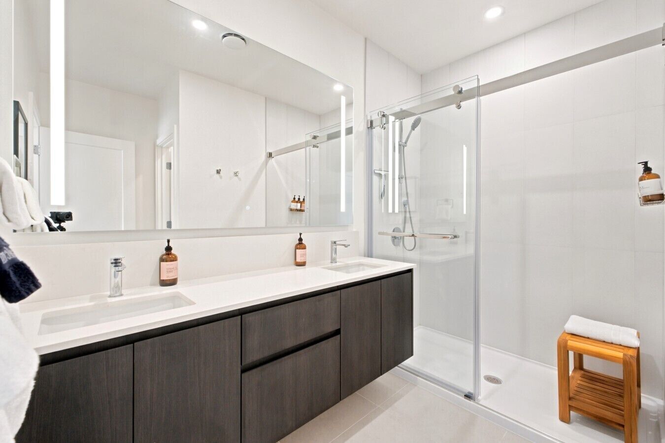 Une salle de bain moderne avec un grand miroir, deux lavabos, des meubles sombres, une douche en verre et un petit tabouret en bois.