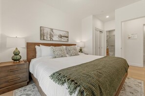 Quarto com cabeceira de madeira, uma cama grande com roupa de cama branca, duas almofadas estampadas, um cobertor verde, uma mesa de cabeceira com abajur e um quadro na parede.