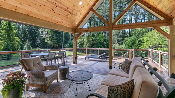 Una terraza techada al aire libre con vigas de madera, un sofá, sillas, una mesa y vistas a una zona boscosa.