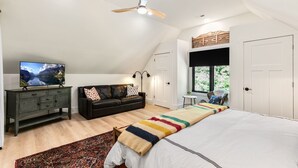 Un dormitorio con una cama grande, un televisor en un mueble, un ventilador de techo y una ventana con vistas a vegetación.