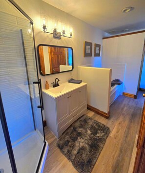 A bathroom with a glass shower enclosure, a white sink with a black faucet, a mirror with a wooden frame, and a small rug on the floor.