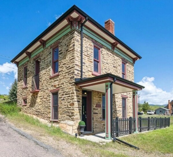 A stone building with a gabled roof, a front porch, and a black metal fence.