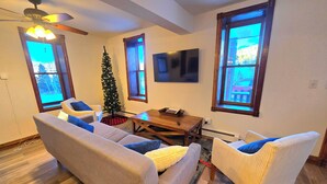 Un salon avec un canapé, des fauteuils, une table basse, un petit sapin de Noël et un téléviseur fixé au mur.