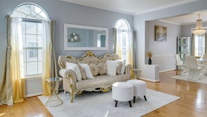 A spacious living room with a large, ornate sofa, a white ottoman, and a glass coffee table. - 'The Mirror' - Elegant & Dog-Friendly DMV Home (Upper Marlboro)