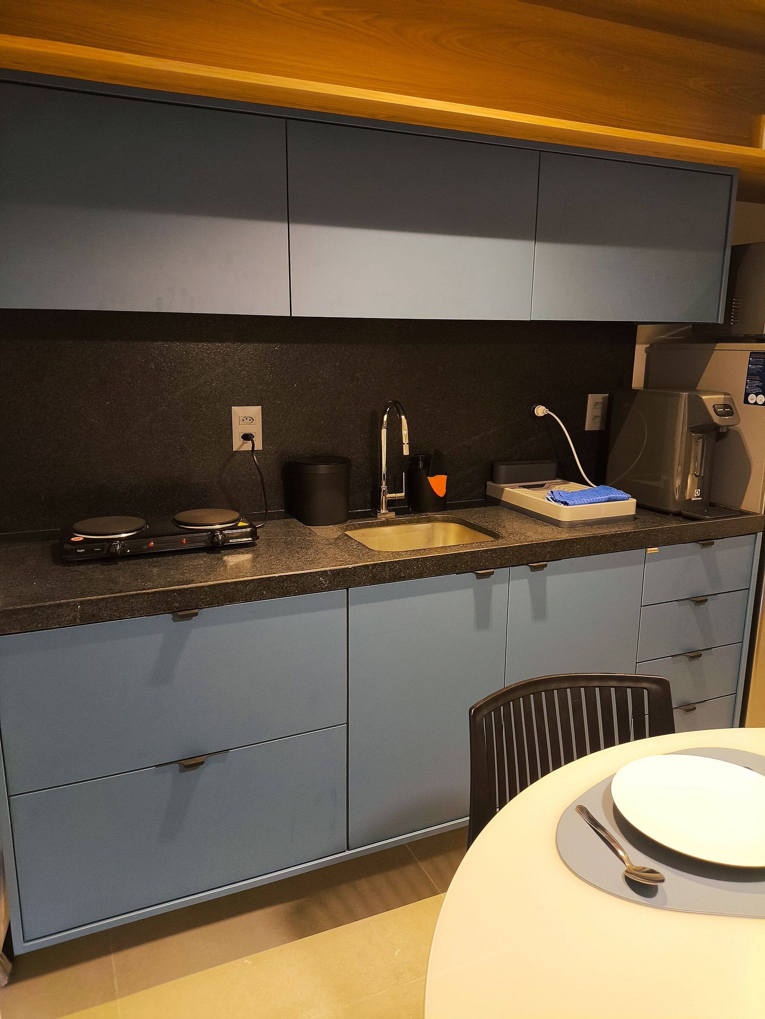 A compact kitchen with a sink, stove, and microwave. There is a chair and a table with a plate and a spoon.