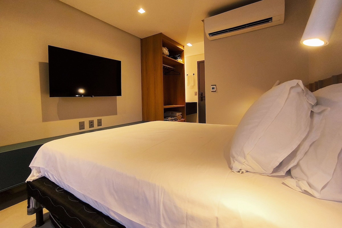 A hotel room with a bed, a TV mounted on the wall, an air conditioning unit, and a closet with hanging clothes.