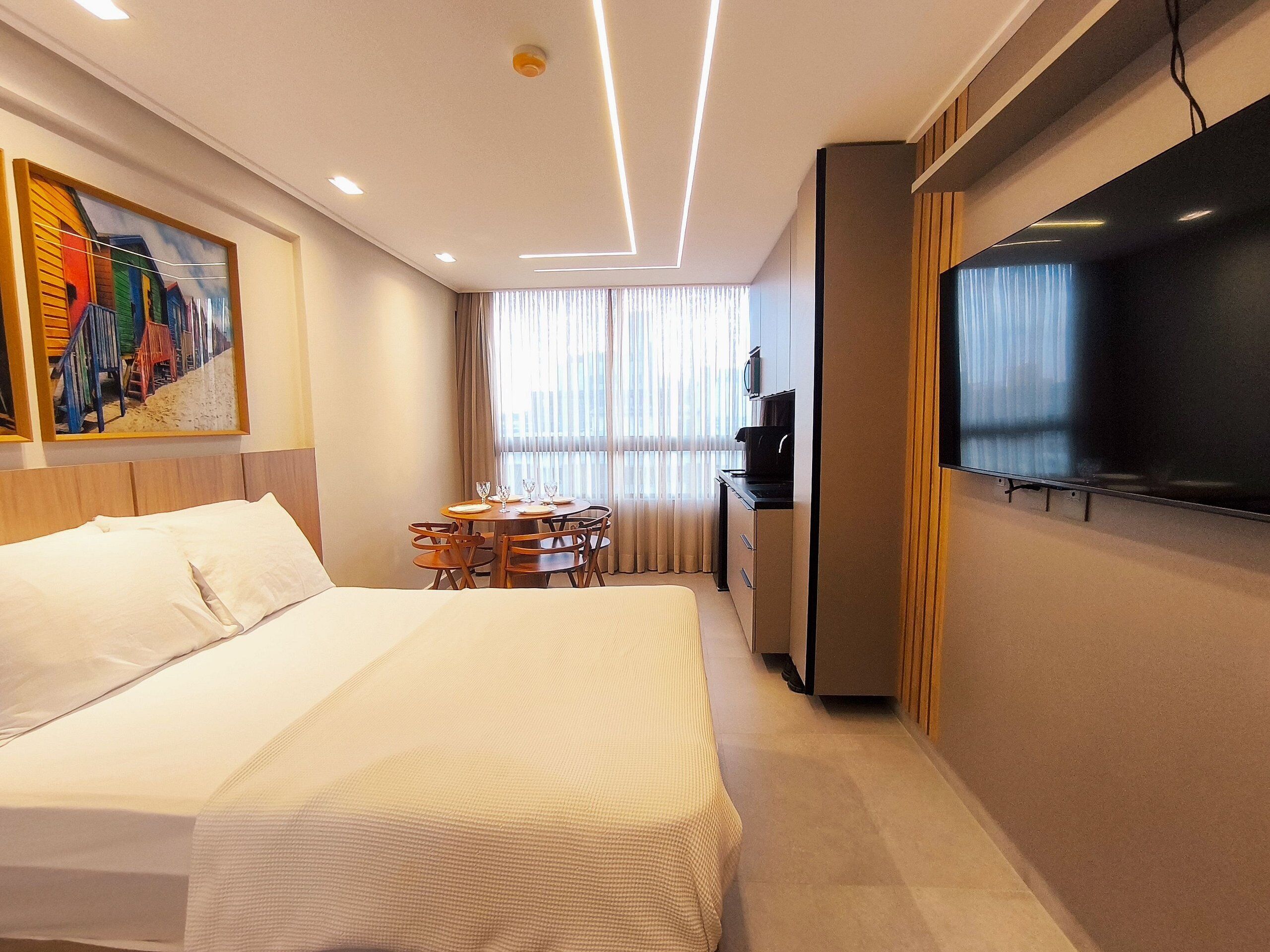 A hotel room with a bed, a dining table with chairs, a TV, and a painting on the wall.