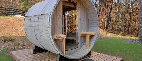 Estructura cilíndrica de madera con entrada abierta, un pequeño rincón de estar en su interior y una terraza de madera al frente.