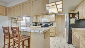 A kitchen with wooden cabinets, a central island with bar stools, a microwave, and a refrigerator. - Golf, Fish & More! Peaceful Home in Helendale (Helendale)