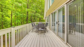 A deck with a table and chairs, surrounded by glass doors and a white railing, overlooking a forested area.