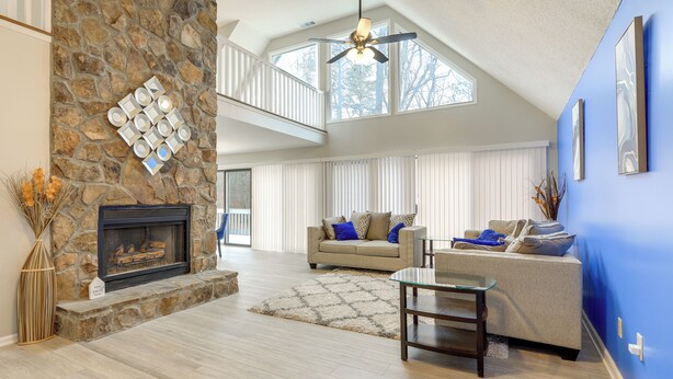 A spacious living room with a stone fireplace, a ceiling fan, and a large window providing natural light.