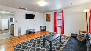 Una sala de estar con piso de madera, un sofá negro, un tapete estampado, una mesita y un televisor montado en la pared.